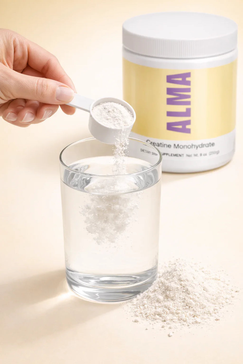 Hand pouring ALMA creatine monohydrate powder into glass of water with supplement container and powder pile